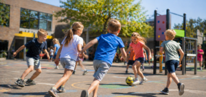 Group of children playing soccer on a sunny schoolyard, chasing a yellow ball with a climbing structure in the background.
