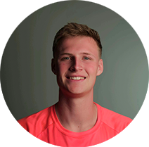 Smiling young man with short light brown hair wearing a coral athletic shirt, posed against a green-gray studio backdrop.