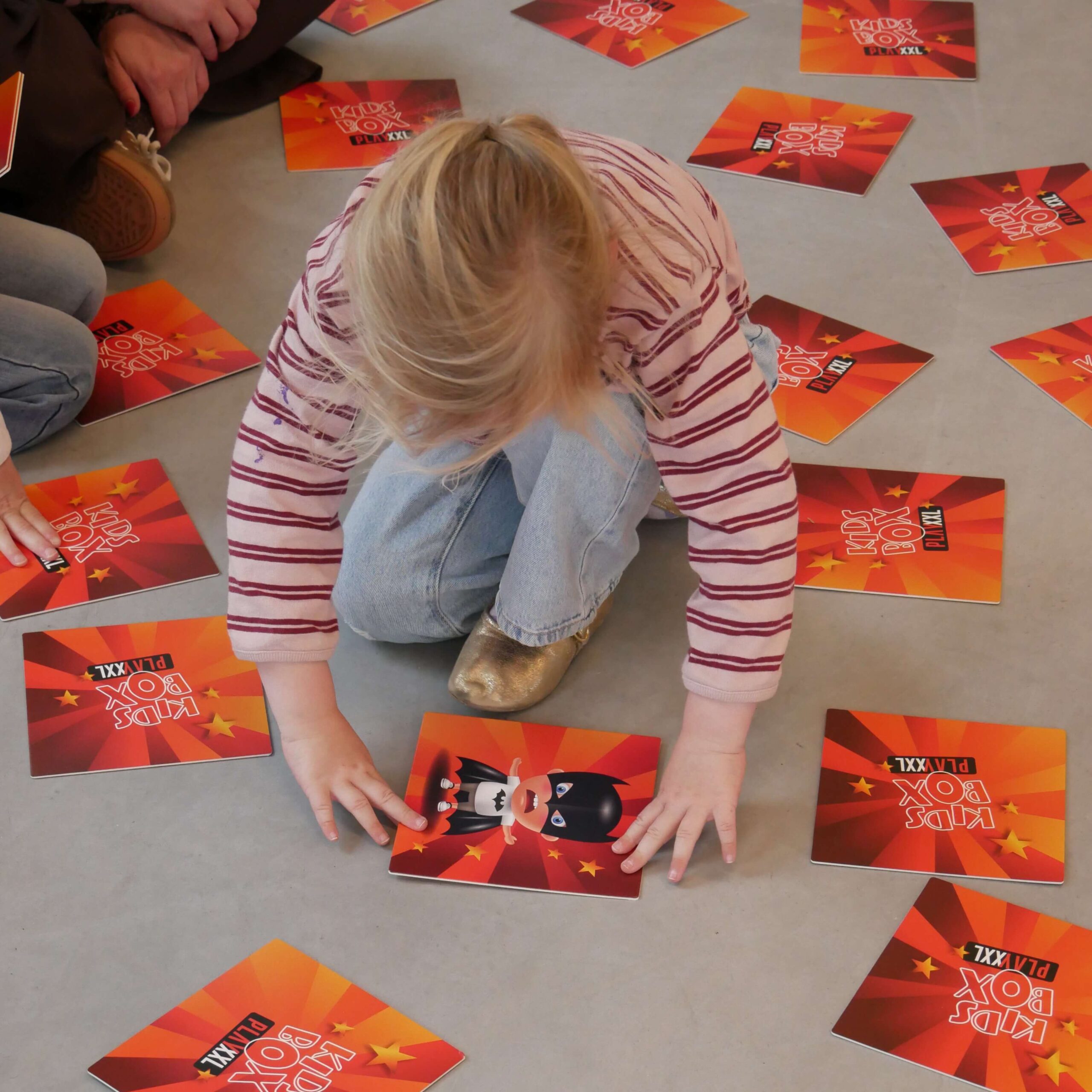 Top-down view of a child sitting on a gray floor, surrounded by orange-red cards that say 'Kids Box' with star graphics. The child wears a striped shirt and denim pants.