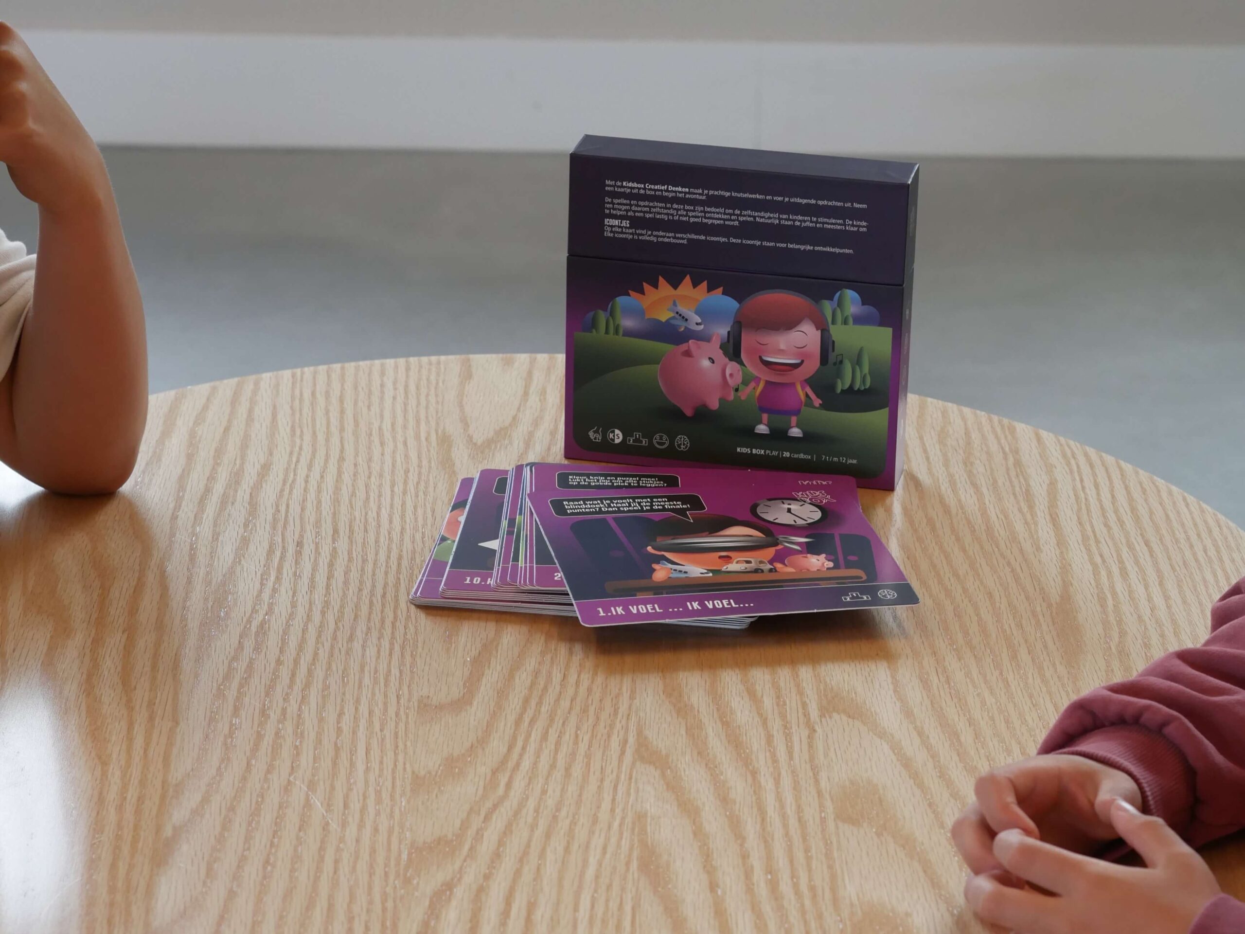 Colorful children's game box and cards spread on a wooden table, with a child's arm visible at the left edge of the image.
