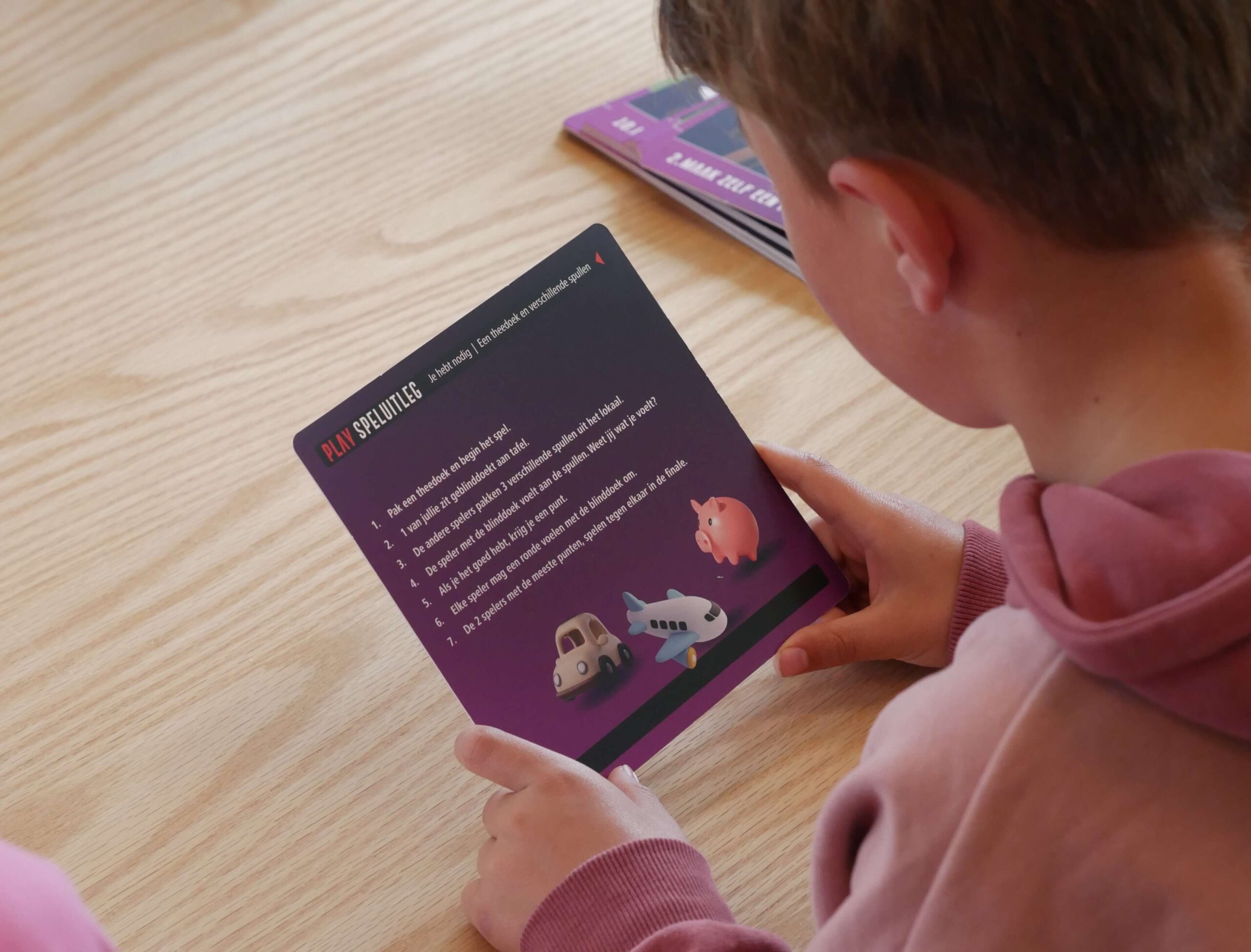 Child holding and reading a purple booklet with cartoon vehicle illustrations on a wooden table.