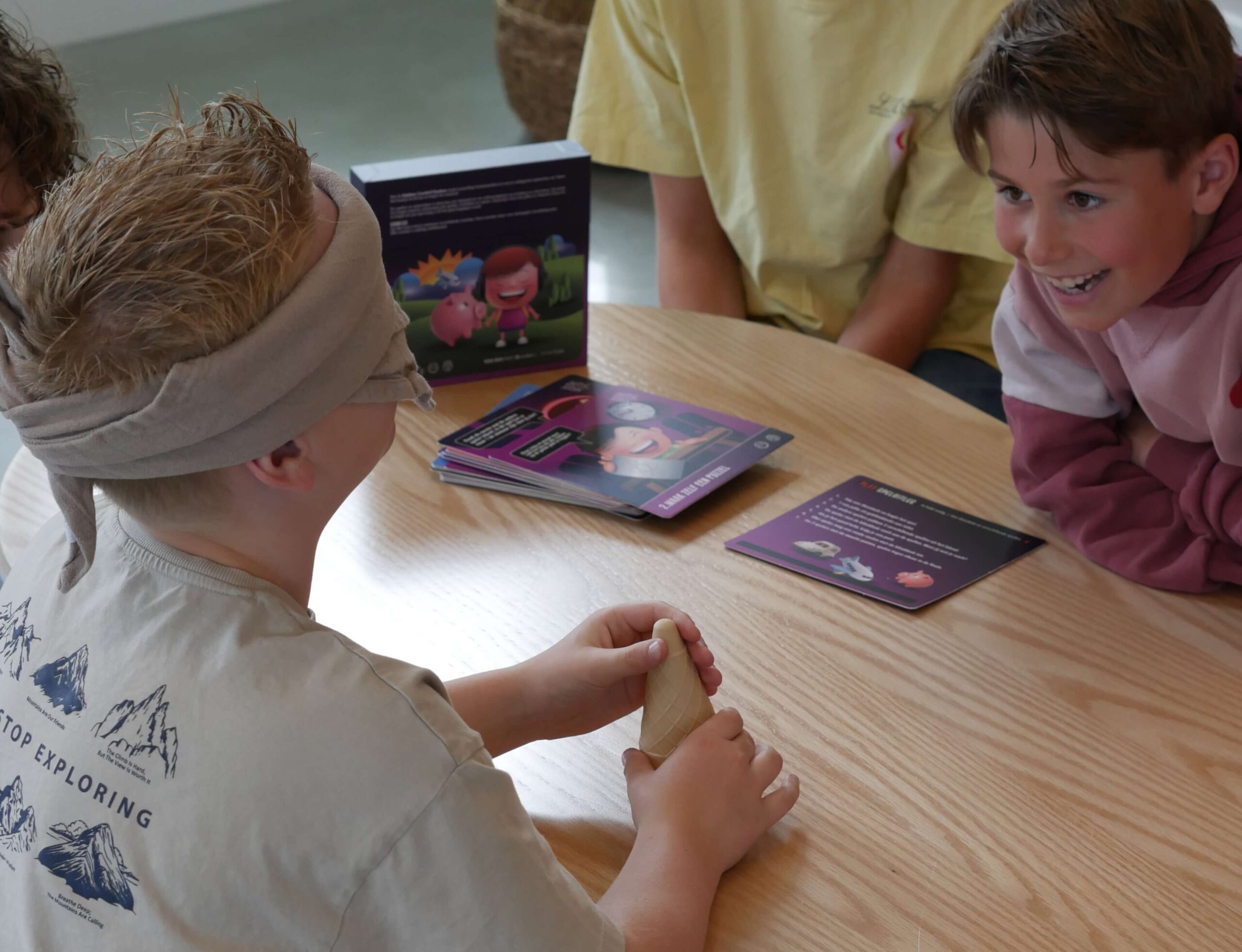 Children seated at a wooden table, playing a card/board game with a purple illustrated box and colorful cards on the table.