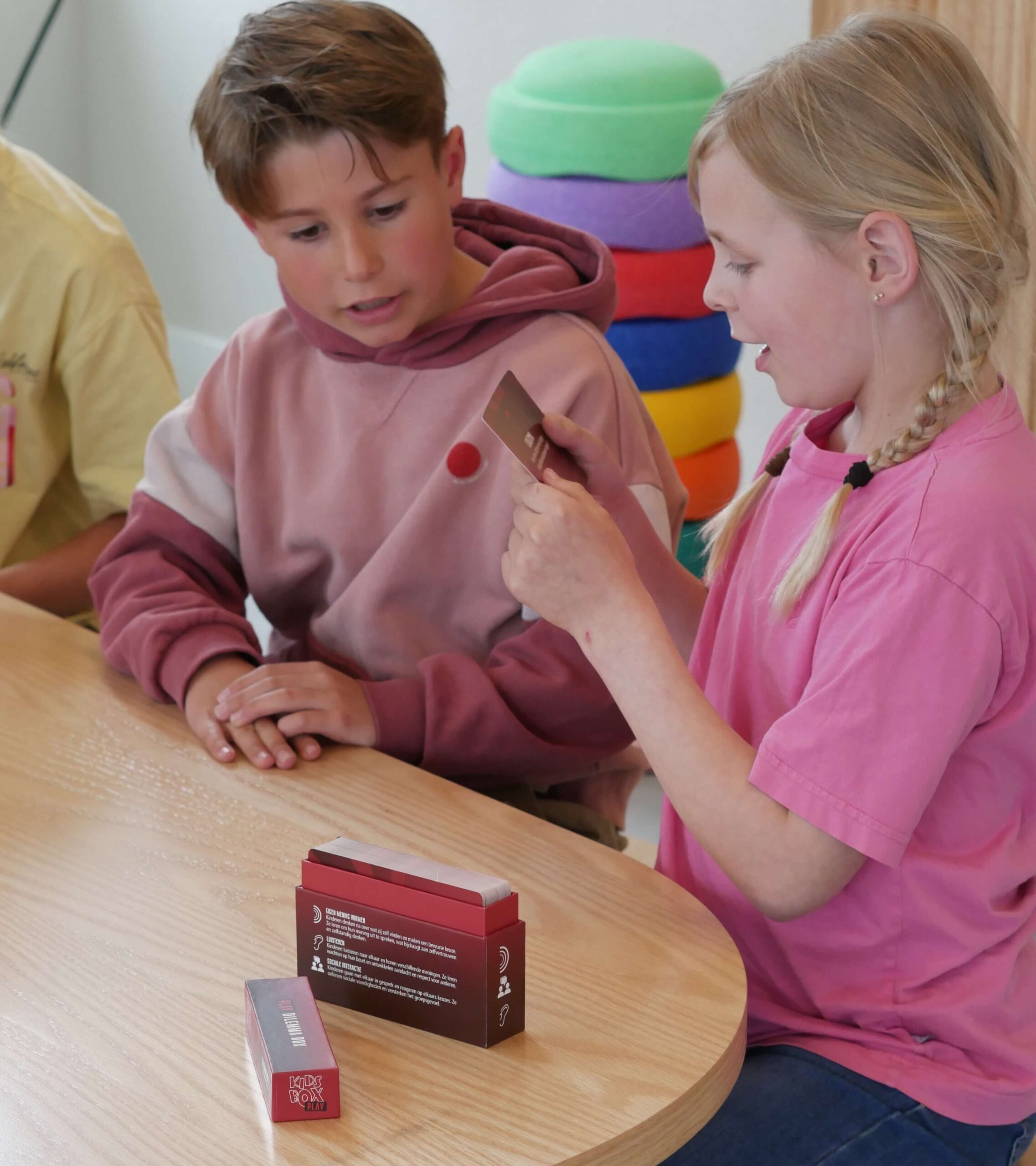 Two children sit at a wooden table with small red game boxes in front of them, chatting while handling a card or box piece.