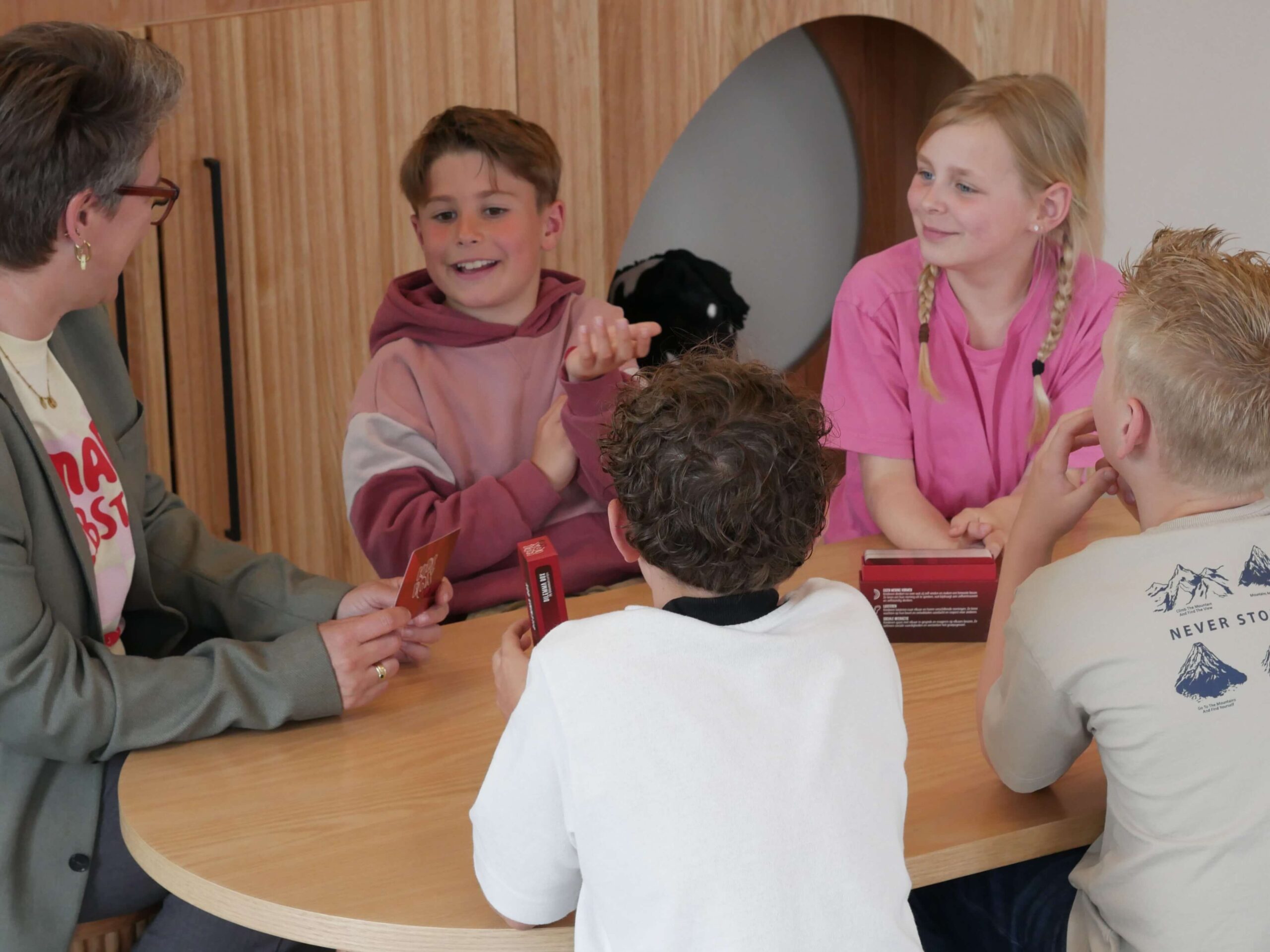 Adult with glasses and a gray blazer talks with a group of children around a wooden table, holding red cards.